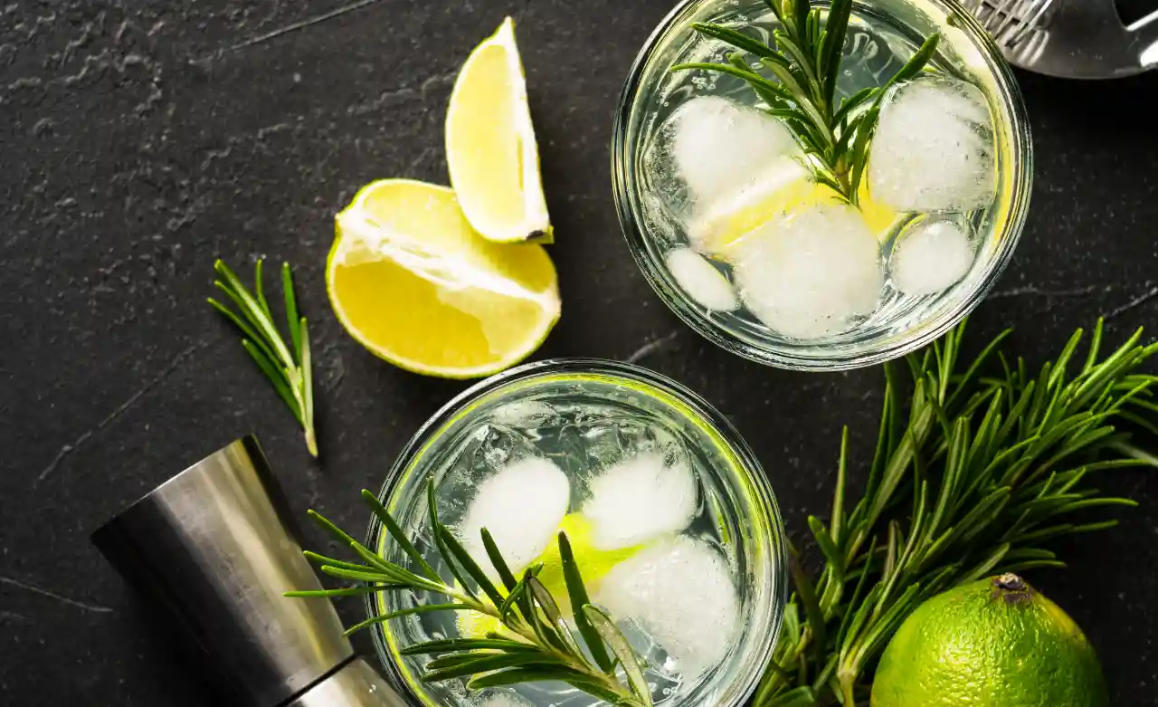 Zwei Gin Tonic mit Eis, Rosmarin und Limette auf dunklem Stein, daneben ein BarmaĂź.