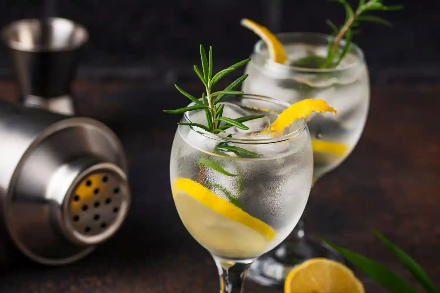 Gin Tonic im Ballonglas mit Eis, Rosmarin und Zitronenzeste, dahinter ein zweites Glas und ein Shaker.